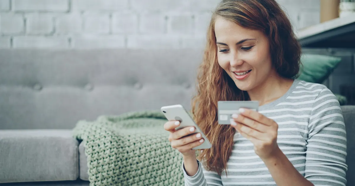 Kanepede oturan genç kadın telefon ve banka kartıyla online ödeme yapıyor